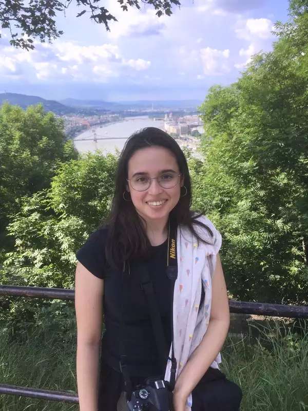 Photo of Ada smiling in front of a railing overlooking a city (with trees and a body of water also visible)