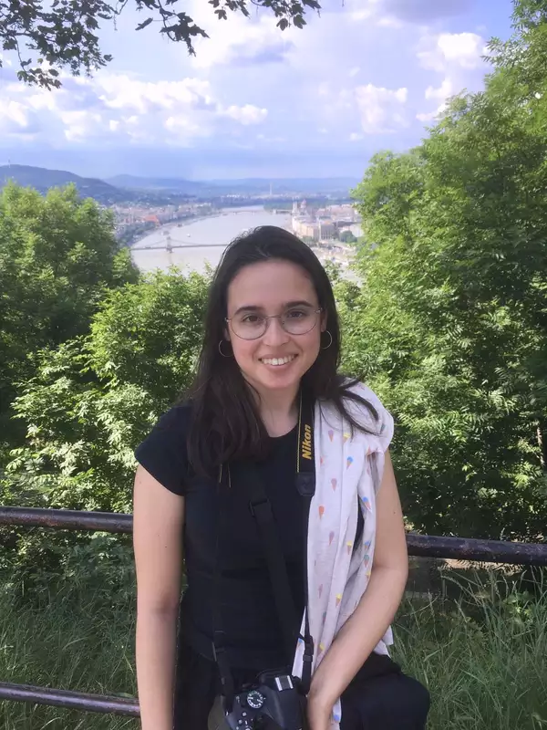 Photo of Ada smiling in front of a railing overlooking a city (with trees and a body of water also visible)