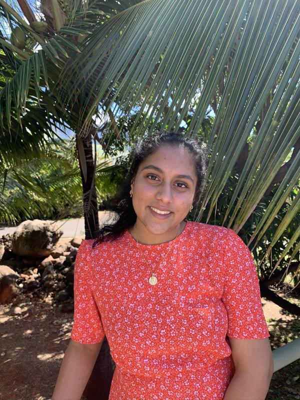 Photo of Ramya in a red shirt with flowers in front of a tree.